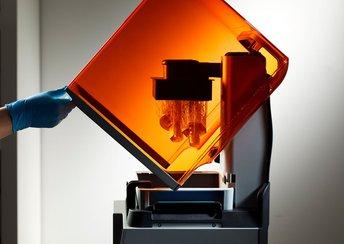 a blue-gloved hand lifts open the orange lid of a Form 4 3D printer