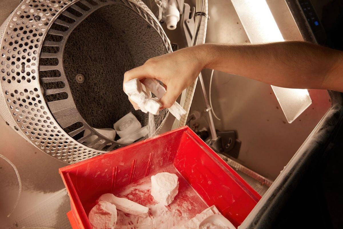a hand picks up powdery white sls 3D printed parts out of a red bucket and places them in the automated tumbling basket of the Formlabs Fuse Blast