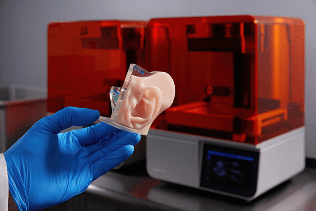 3D printed model of an ear being held in a gloved hand in front of two Form 4B 3D printers