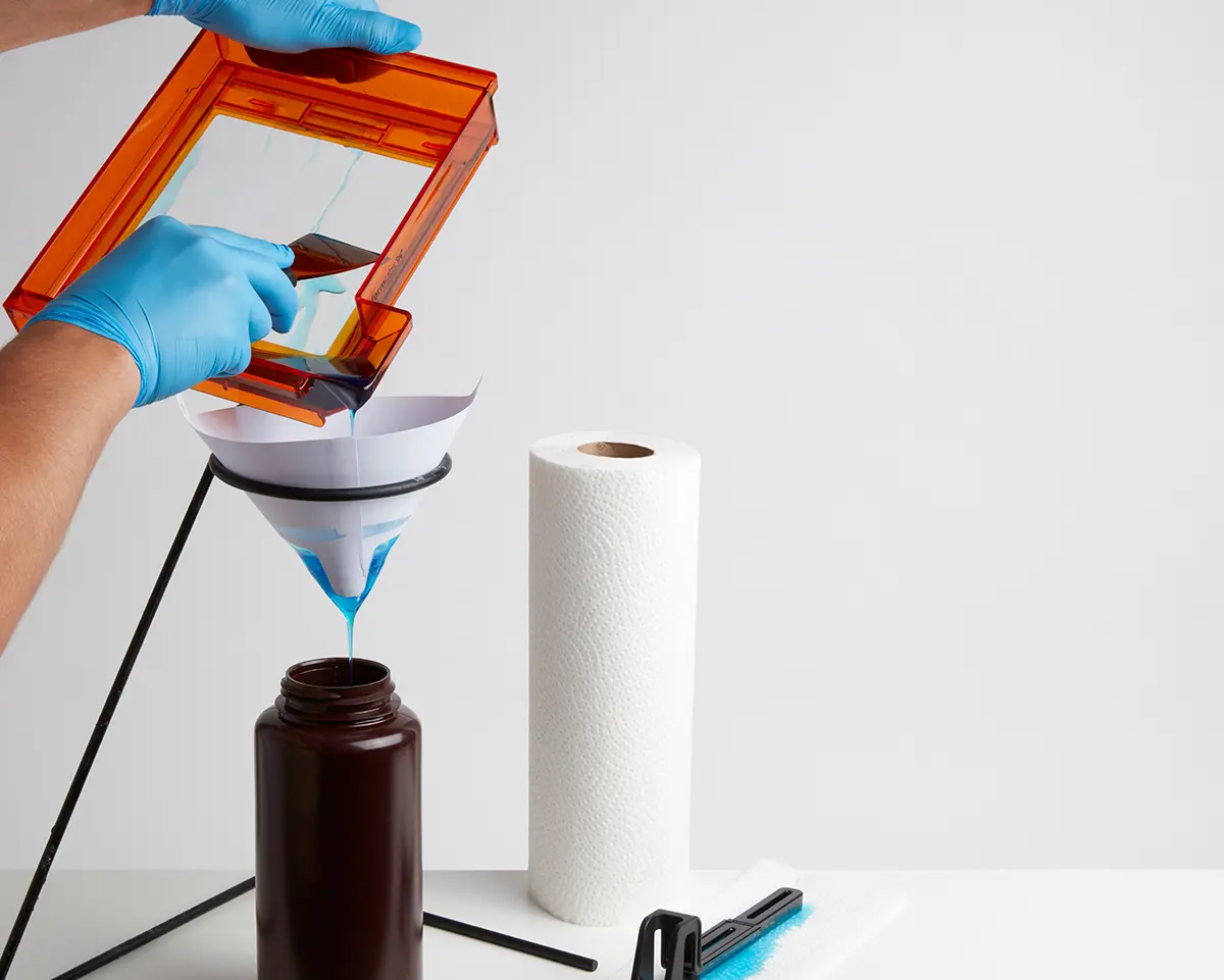 Form 2のレジンタンク内のレジンをヘラで掬い取って、不透明なプラスチックの容器の上に吊り下げられた塗料フィルタの上に移します。