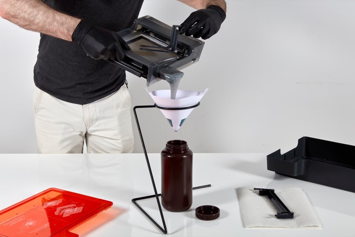 Form 3のレジンタンク内のレジンをヘラで掬い取って、不透明なプラスチックの容器の上に吊り下げられた塗料フィルタの上に移します。