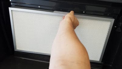 The grate has been removed. Someone is reaching out to grip the side of the vent filter