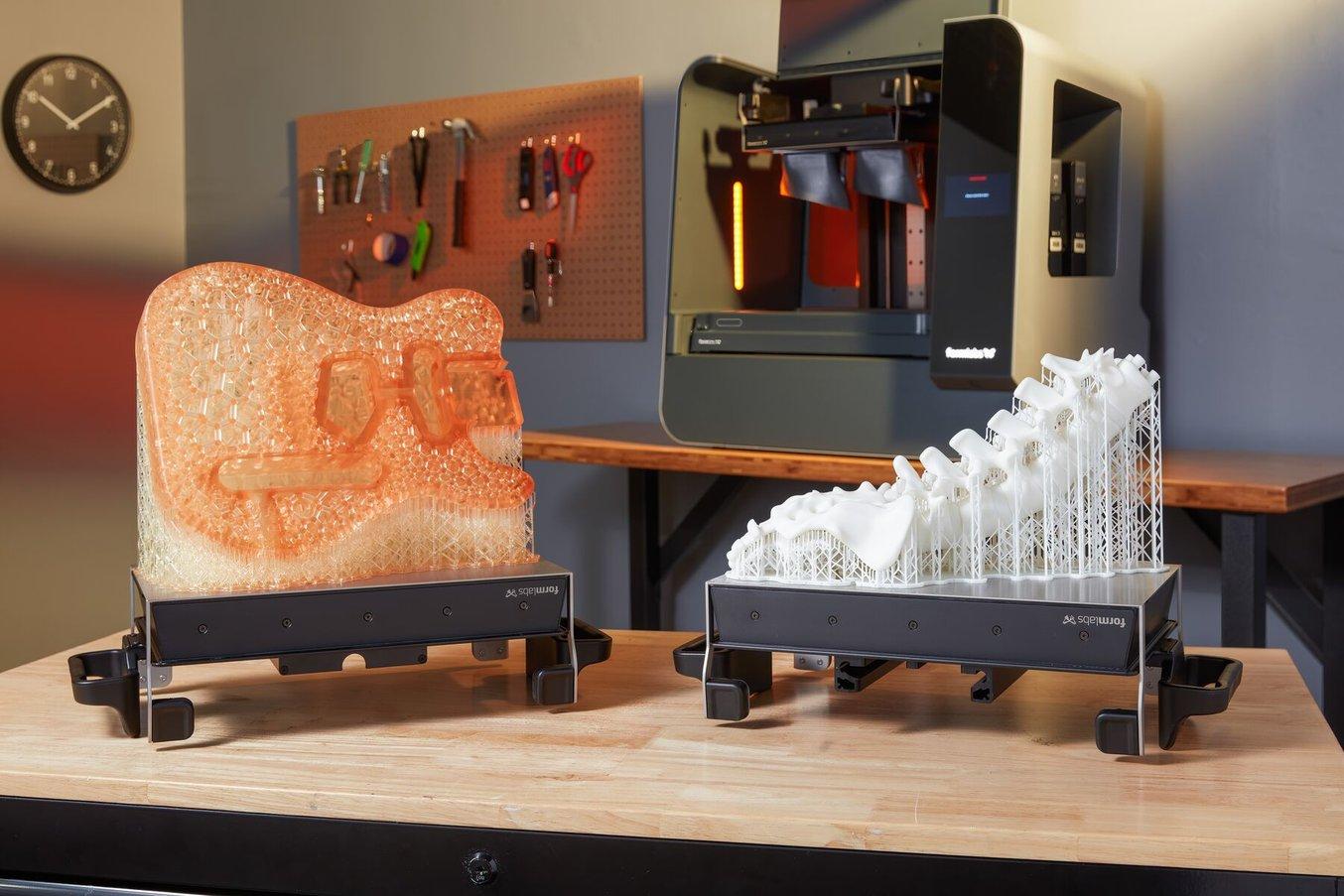 Un cadre de guitare imprimé en 3D et un modèle anatomique de la colonne vertébrale imprimé en 3D sur des plateformes de fabrication sont posés sur une table devant une imprimante 3D stéréolithographique (SLA) Form 3L.