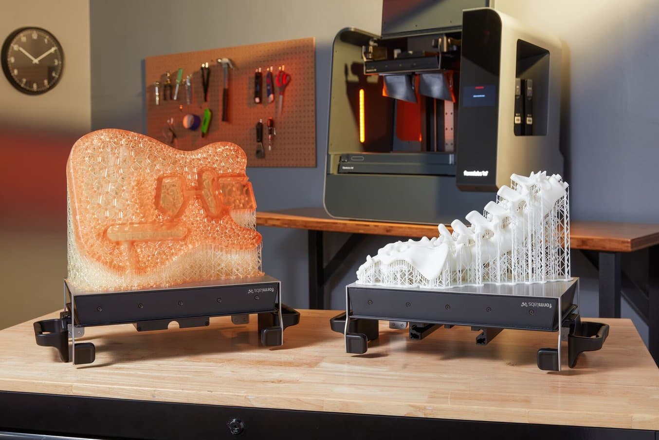 Un cadre de guitare imprimé en 3D et un modèle anatomique de la colonne vertébrale imprimé en 3D sur des plateformes de fabrication sont posés sur une table devant une imprimante 3D stéréolithographique (SLA) Form 3L.