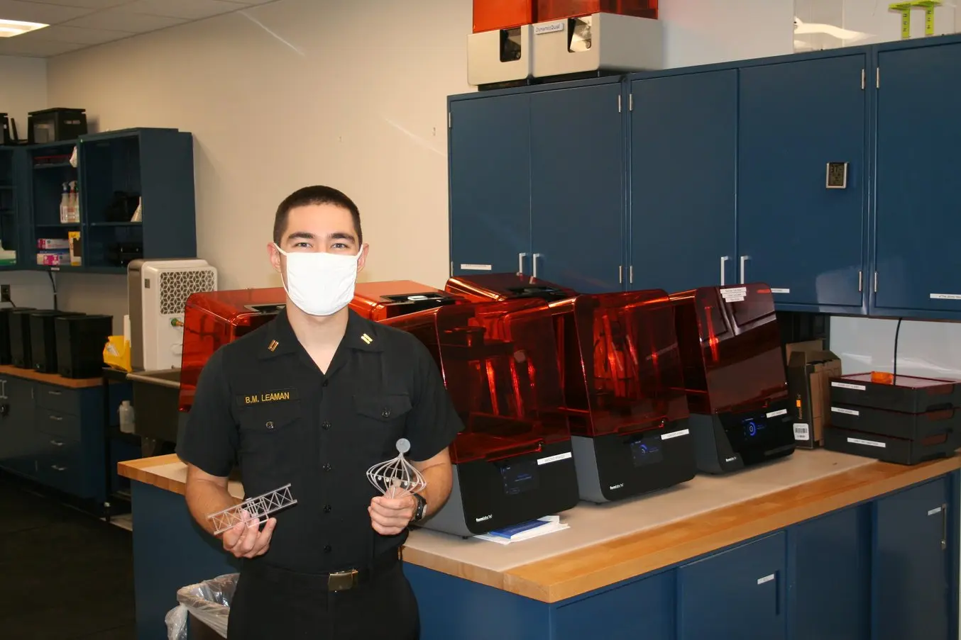 A young man holds intricate 3D printed models and stands in front of six Form 3 3D printers in a workshop.