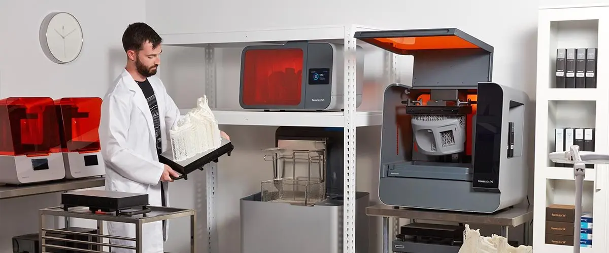 A person in a white lab coat inspects a 3D printed model of a human spine in front of several 3D printers.