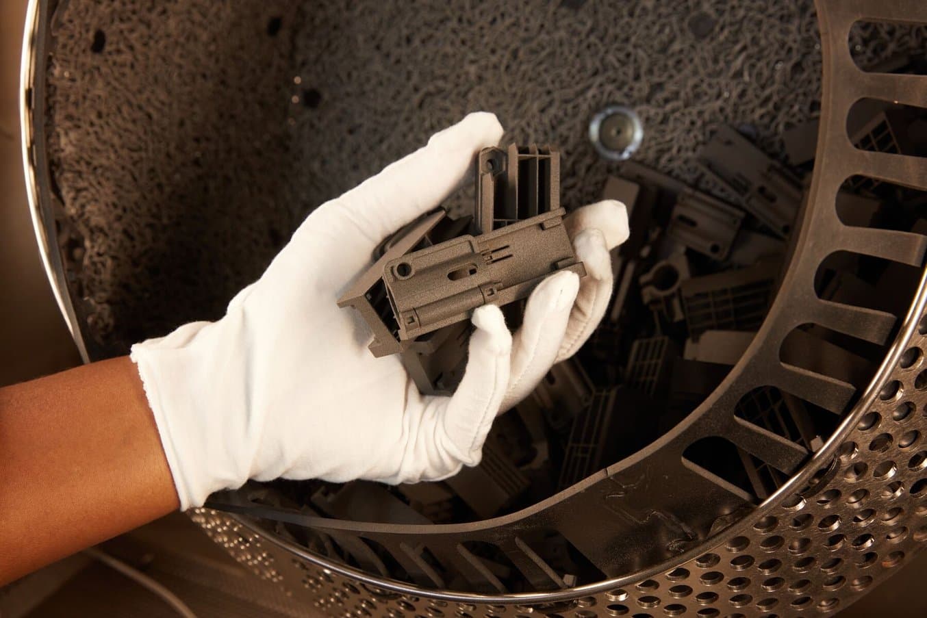 a white gloved hand holds SLS 3D printed parts in front of the tumbling basket of the Fuse Blast