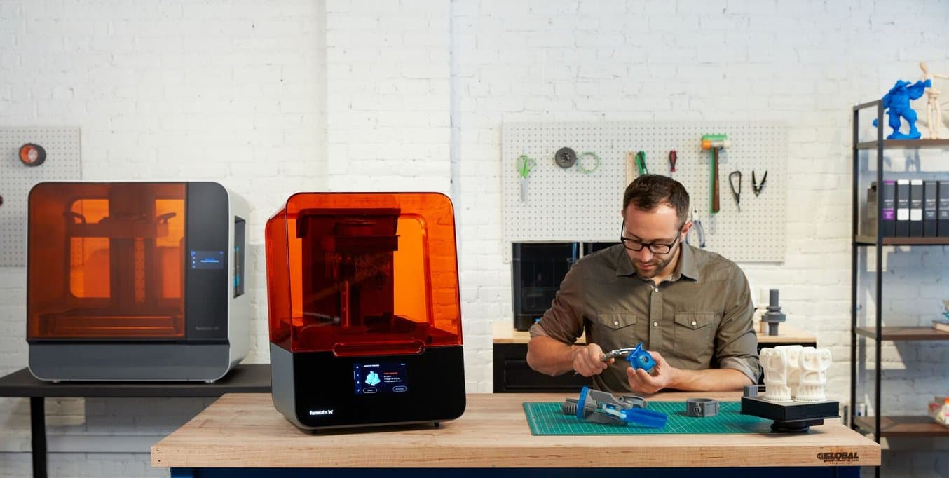 An engineer measures a 3D printed part. He sits next to a Form 3 and in front of a Form 3L.