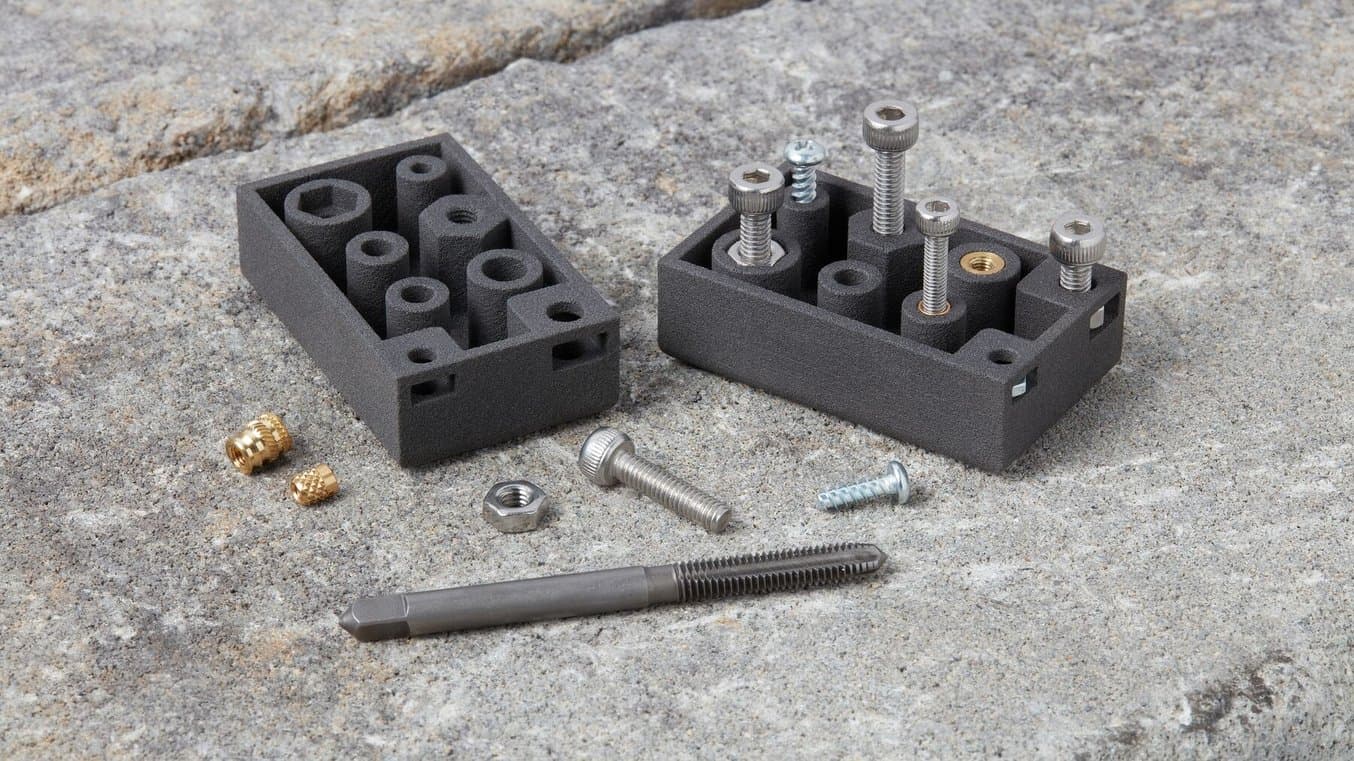 Two 3D printed test parts, plus screws, taps, and inserts scattered on a concrete background.