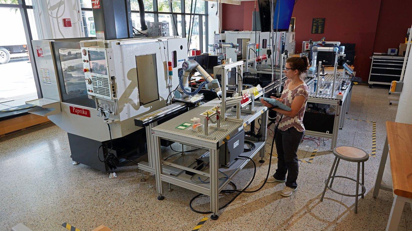 A woman operates a pick-and-place robotic arm using a tablet. Milling machines and other equipment are visible nearby.