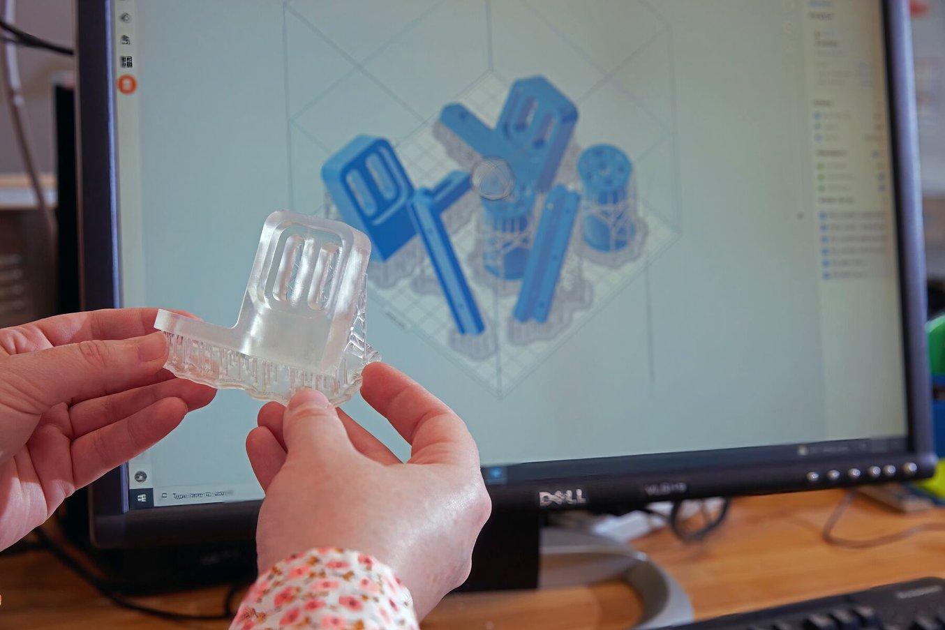 A person’s hands hold a translucent 3D-printed part, in front of a computer showing the part and several others as set up for printing.