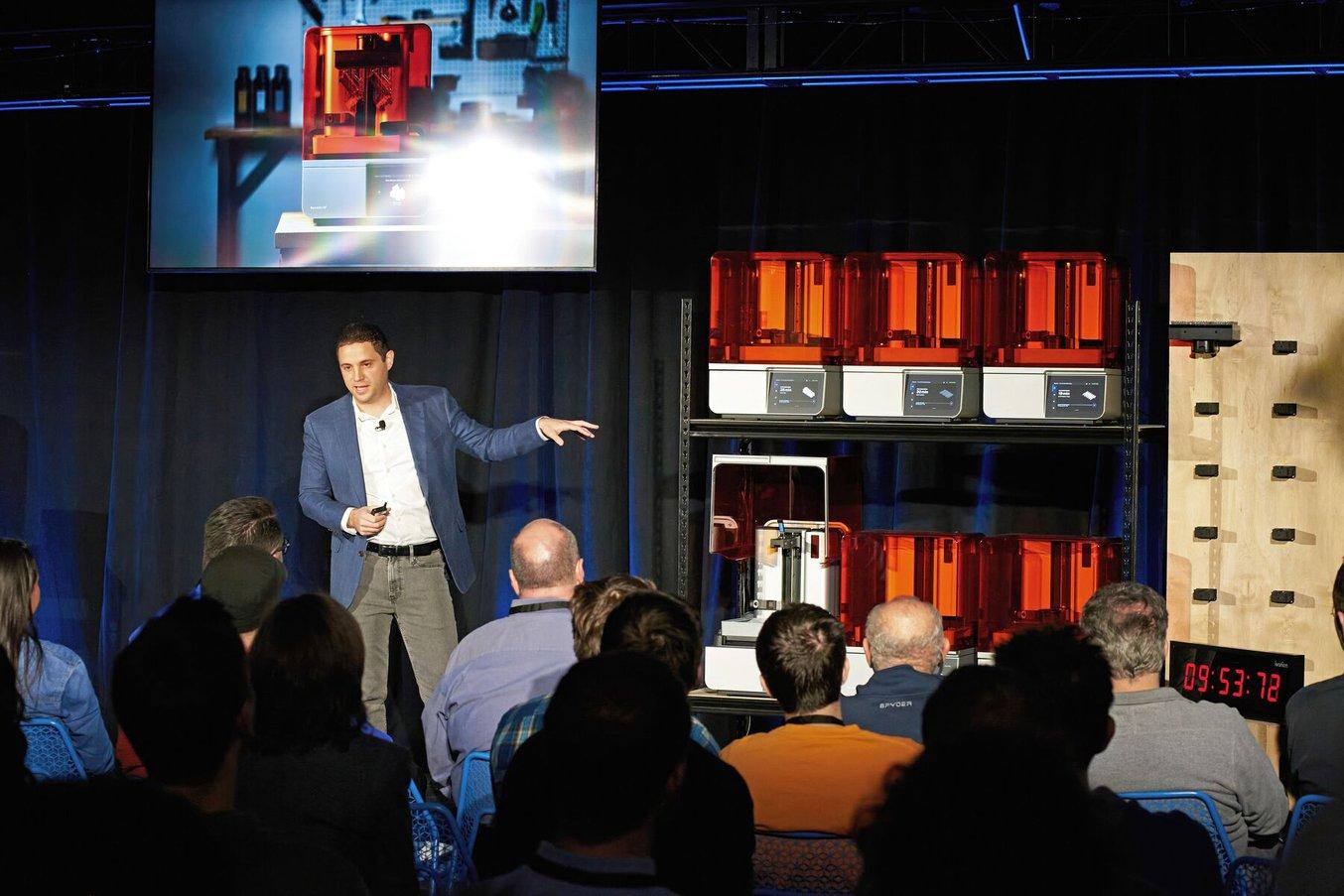 a man stands in front of a fleet of 3D printers while giving a presentation to a crowd