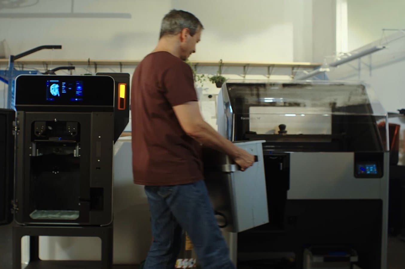 A person moves a build chamber from a Fuse 1 3D printer to a Fuse Sift powder recovery station.