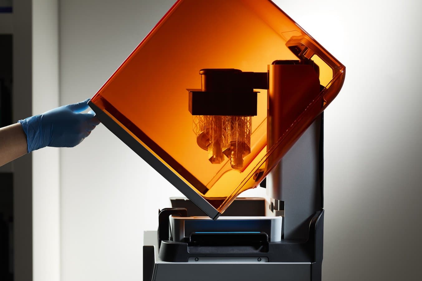 a blue-gloved hand lifts the orange cover of a Form 4 3D printer