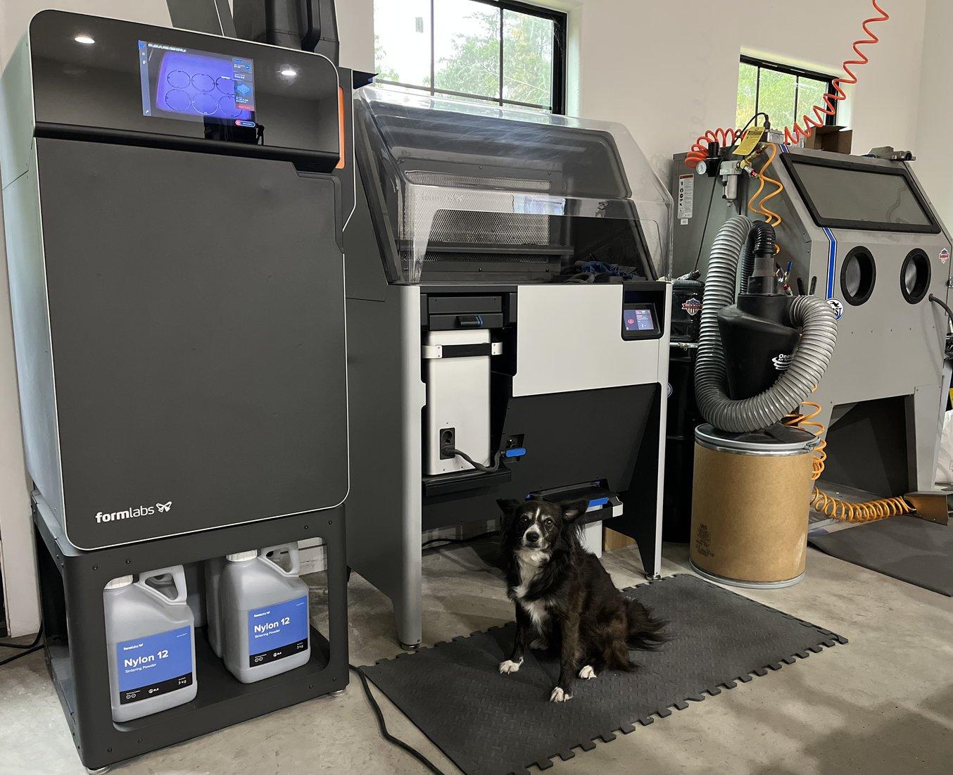 Der 3D-Drucker Fuse 1 für selektives Lasersintern (SLS), die Nachbearbeitungseinheit Fuse Sift, ein Medienblaster und ein Hund.