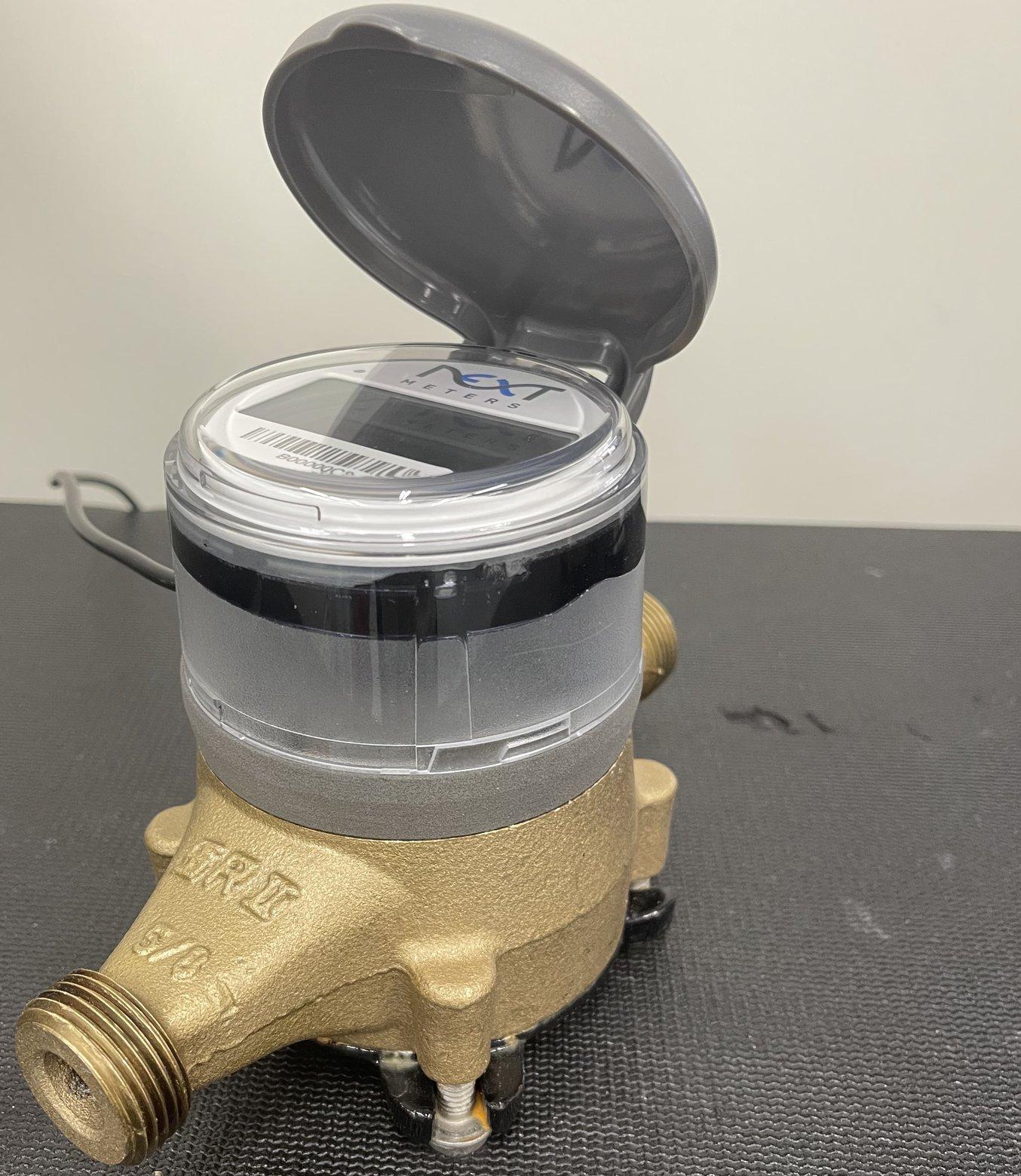 A water meter (clear casing) fitted onto a unique water system spigot (gold plated) via a custom-printed Fuse 1 SLS fixture (grey middle part).