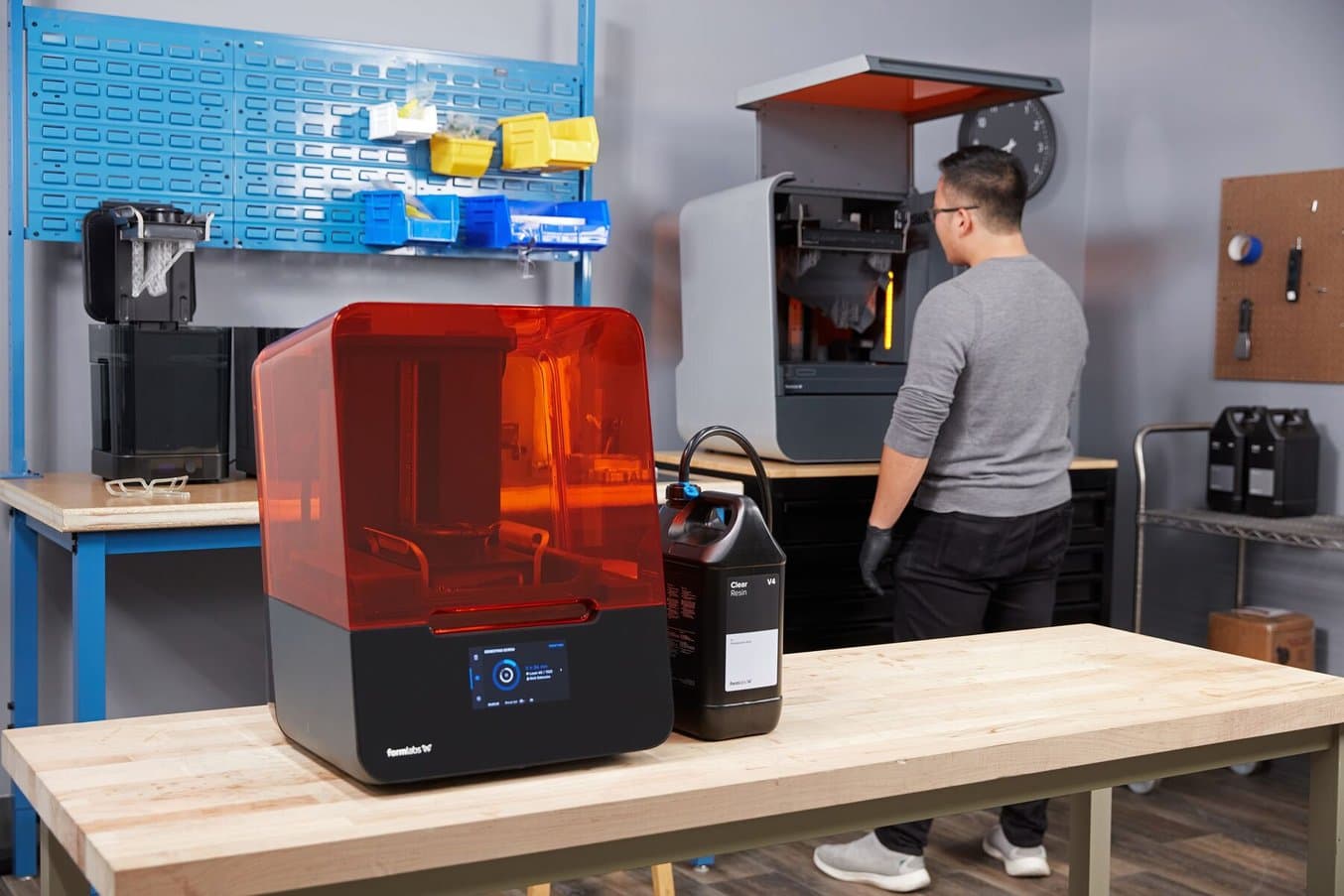 the Form 3+ and Resin Pumping System in the foreground on a table, a man stands at the Form 3L in the background