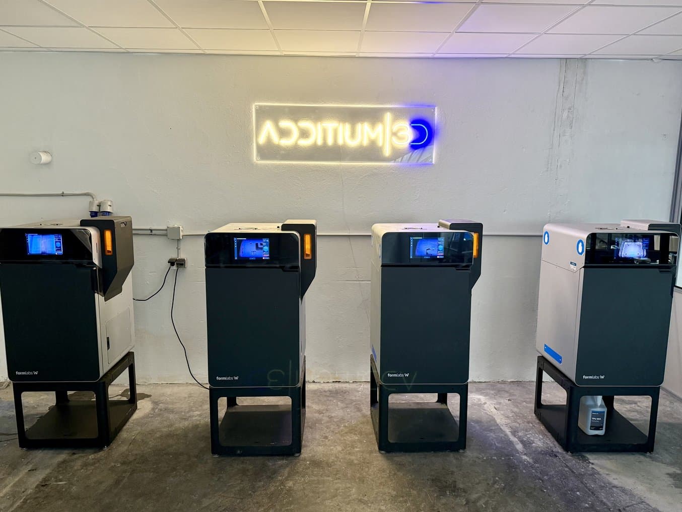 Vier Fuse 1+ 30W SLS-3D-Drucker unter einem Neonschild mit der Aufschrift „Additium3D“.