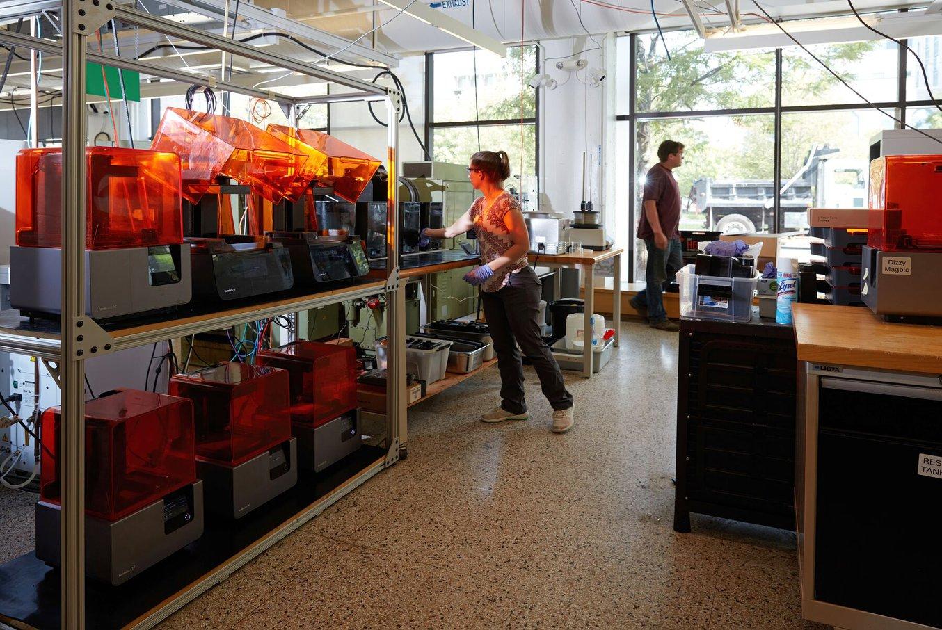A woman uses equipment next to a rack of six 3D printers.