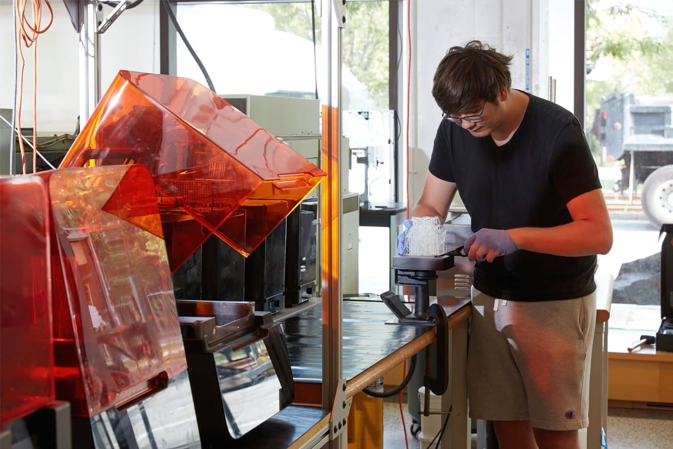 A student uses a prying tool to carefully remove a 3D printed part from a build platform, which is held in place by a clamped jig.