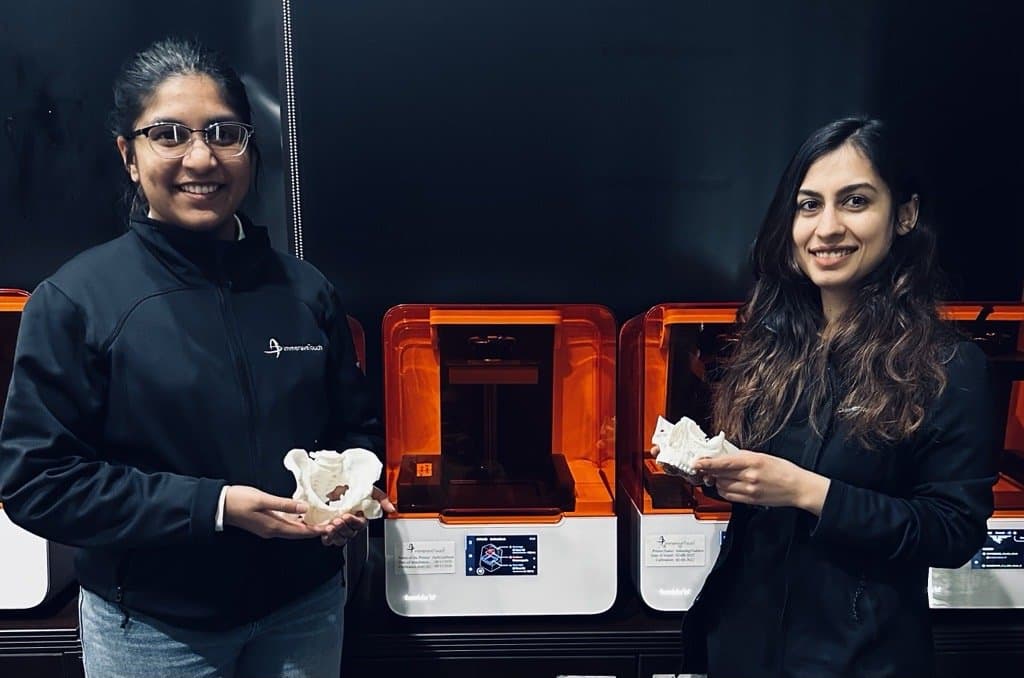 Two women holding anatomical models stand in front of Form 3B+ 3D printers