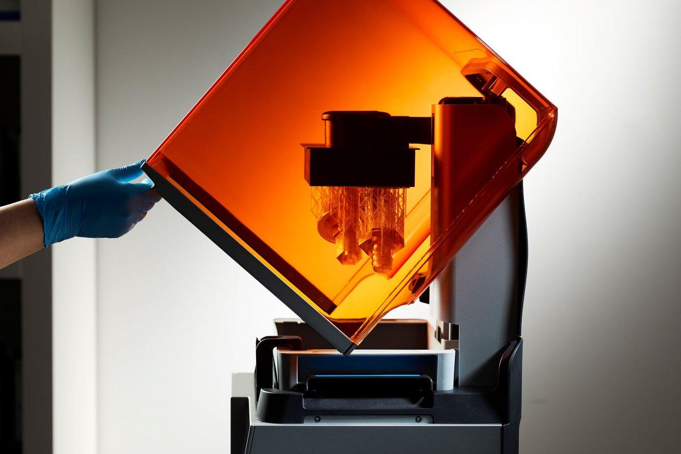 a blue-gloved hand lifts the orange cover of a Form 4 3D printer