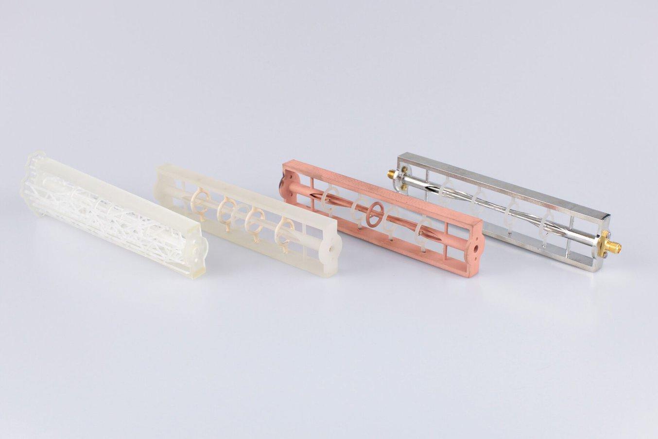 Four versions of the same design, laid out on a gray background. From left to right: 3D printed clear part on supports, 3D printed clear part with support material removed, part plated in copper, and part plated in tin.
