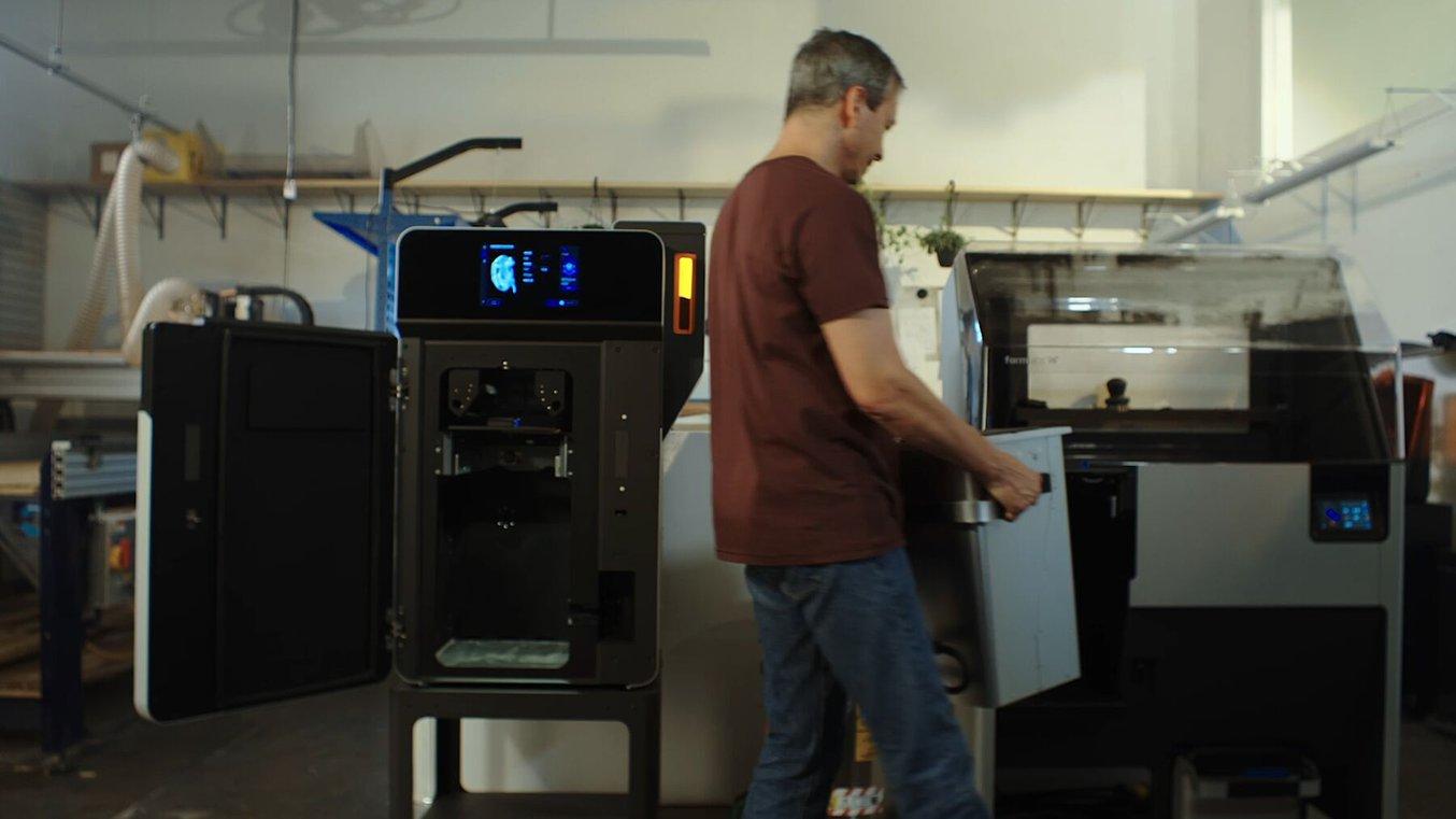 A person moves a build chamber from a Fuse 1 3D printer to a Fuse Sift powder recovery station.